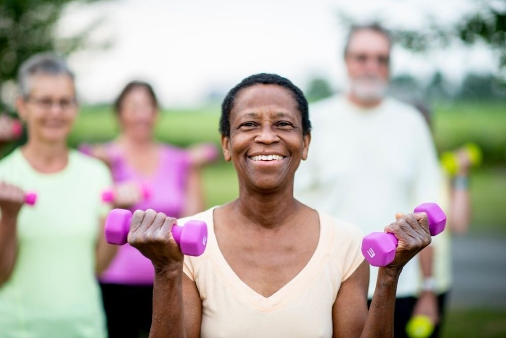 An older woman smiling as she lifts a 3kg dumbbell in each hand in a fitness class.